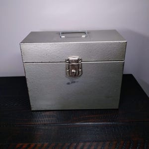 May include: A rectangular, silver-colored metal storage box with a handle on top and a latch closure. The box is sitting on a dark wooden surface.