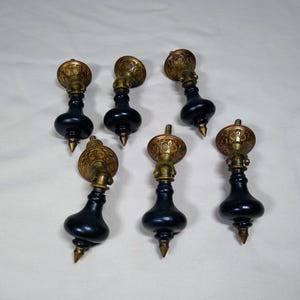 May include: A set of six drawer pulls featuring black, teardrop-shaped wooden bodies with pointed tips. Each pull has a circular, ornate brass mounting plate with intricate floral engravings and a hinged brass connector at the top.