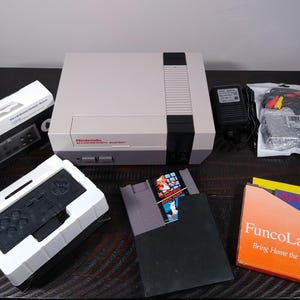 May include: A Nintendo Entertainment System console with accessories, including a controller, game cartridges, and power adapter. The console is light grey with a black stripe and red lettering. The game cartridges are in black cases, and one box reads "FuncoLand".