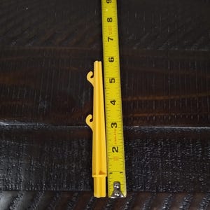May include: A yellow plastic tent stake with three prongs at the top and a pointed end. The stake is measured against a tape measure, showing it is approximately 5 inches long.