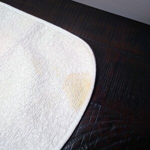 May include: A white, rounded-corner, absorbent cloth with a yellow stain in the corner. The cloth has a textured surface and is set against a dark brown wooden surface.