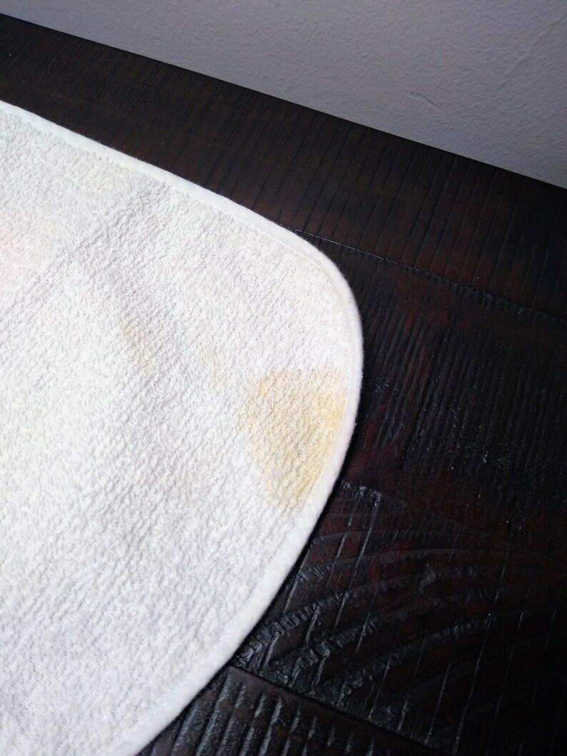 May include: A white, rounded-corner, absorbent cloth with a yellow stain in the corner. The cloth has a textured surface and is set against a dark brown wooden surface.