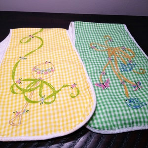 May include: Two baby bibs with embroidered designs. One bib is yellow gingham with a rattle design, the other is green gingham with a butterfly and bird design. Both bibs have white terry cloth backing.
