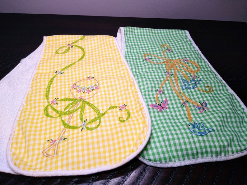 May include: Two baby bibs with embroidered designs. One bib is yellow gingham with a rattle design, the other is green gingham with a butterfly and bird design. Both bibs have white terry cloth backing.