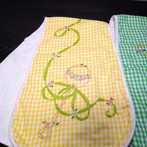 May include: Embroidered baby bibs with a yellow and white gingham pattern. The bibs feature a floral design with green ribbon-like embroidery and small pink and blue flowers. The edges are trimmed with white fabric.