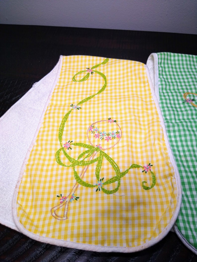 May include: Embroidered baby bibs with a yellow and white gingham pattern. The bibs feature a floral design with green ribbon-like embroidery and small pink and blue flowers. The edges are trimmed with white fabric.