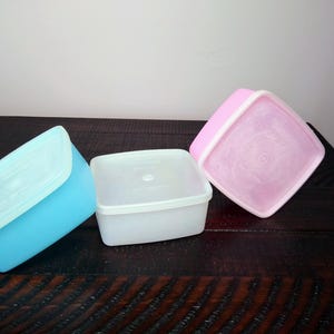 May include: Three square plastic food storage containers in blue, white, and pink. Each container has a matching lid. The containers are on a dark wooden surface.