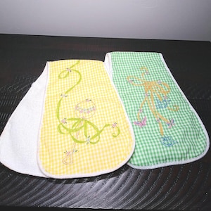 May include: Two baby bibs with a white terry cloth backing. One bib is yellow gingham with a green embroidered design, and the other is green gingham with a tan embroidered design. Both have a floral and butterfly theme.
