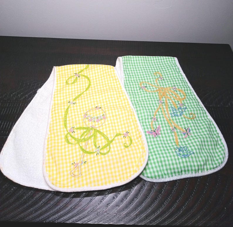 May include: Two baby bibs with a white terry cloth backing. One bib is yellow gingham with a green embroidered design, and the other is green gingham with a tan embroidered design. Both have a floral and butterfly theme.