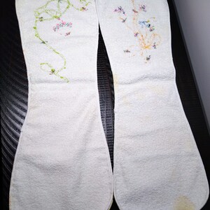 May include: Two white, oblong-shaped cloth items with floral embroidery. One features a green vine with pink flowers, the other has a cluster of blue, pink, and yellow flowers. The items appear to be vintage or handmade.