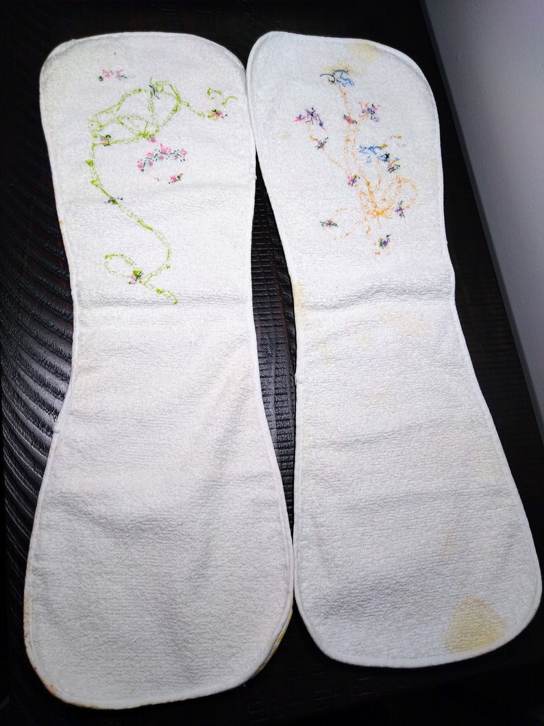 May include: Two white, oblong-shaped cloth items with floral embroidery. One features a green vine with pink flowers, the other has a cluster of blue, pink, and yellow flowers. The items appear to be vintage or handmade.