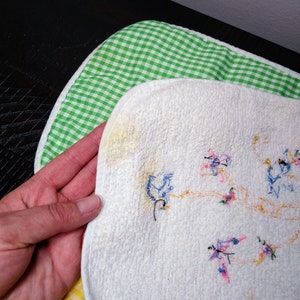 May include: White, embroidered bib with a floral design, held over a green and white checkered bib and a yellow and white checkered bib. The embroidered bib has a textured surface and features blue, pink, and orange thread.