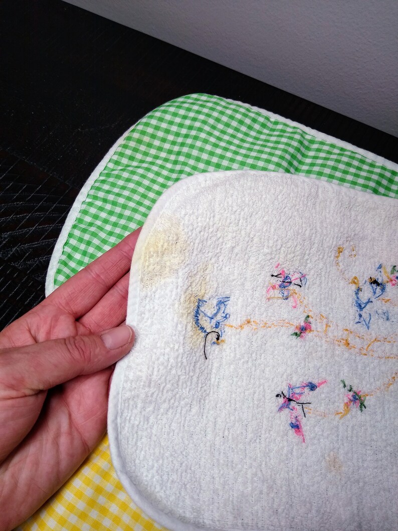 May include: White, embroidered bib with a floral design, held over a green and white checkered bib and a yellow and white checkered bib. The embroidered bib has a textured surface and features blue, pink, and orange thread.