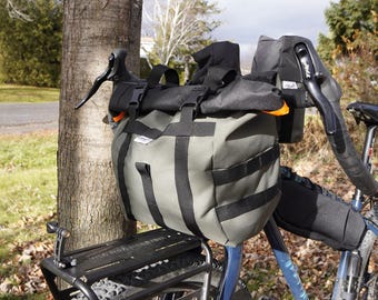 May include: A blue bicycle with a black rack and a green and black canvas bag attached to the rack. The bicycle is parked next to a tree.