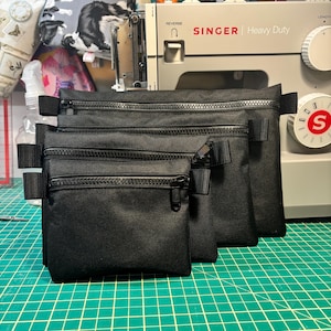 May include: Three black zippered pouches of different sizes, lined up on a green cutting mat. The pouches are made of a durable fabric and have a zipper closure. The pouches are sitting in front of a white Singer sewing machine.