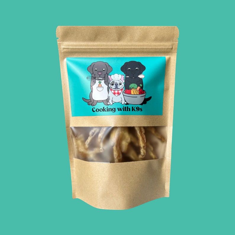 Pu&ograve; includere: Un sacchetto di carta marrone con una finestra trasparente che mostra leccornie per cani a base di zampe di pollo. Il sacchetto ha un'etichetta blu e bianca con un'immagine di cartone animato di tre cani e il testo "Cooking with K9s".