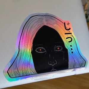 Puede incluir: Pegatina holográfica con un rostro estilizado con rasgos negros, ojos rojos y cabello suelto. El cabello tiene un degradado arcoíris. Hay caracteres japoneses a la derecha. La pegatina está en un portátil.