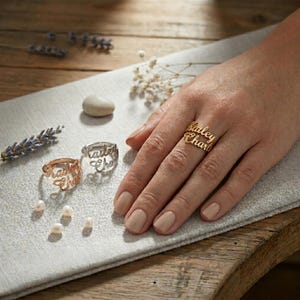 May include: Three personalised name rings in gold, silver, and rose gold are displayed. The rings feature names in a cursive font. Pearls and lavender sprigs are scattered around the rings, adding a delicate touch. The rings are on a neutral surface.