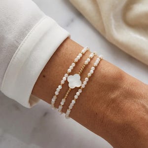 Peut inclure: Un ensemble de trois bracelets de perles composé de petites perles rondes blanches et de perles intercalaires dorées métalliques. Le bracelet central comporte une breloque blanche en forme de trèfle à quatre feuilles.