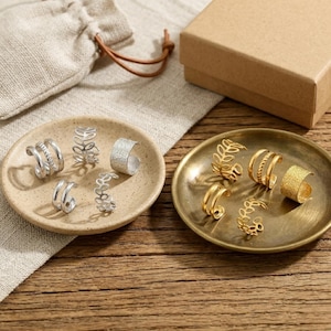 May include: A selection of silver and gold rings on small, round trays. The rings showcase diverse designs, such as textured bands, leaf motifs, and double bands. A small beige drawstring bag and a brown cardboard box are also present.