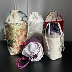 May include: Five drawstring bags made of different fabrics. The bags are in various colors, including white, cream, gray, purple, and red. The bags have different patterns, including floral, geometric, and plain. The bags are all tied with cords and have wooden beads.