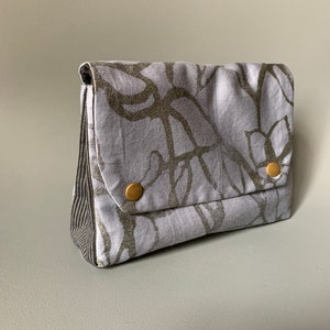 May include: A small, gray fabric pouch with a silver and black abstract pattern. The pouch has a flap closure and two gold buttons.
