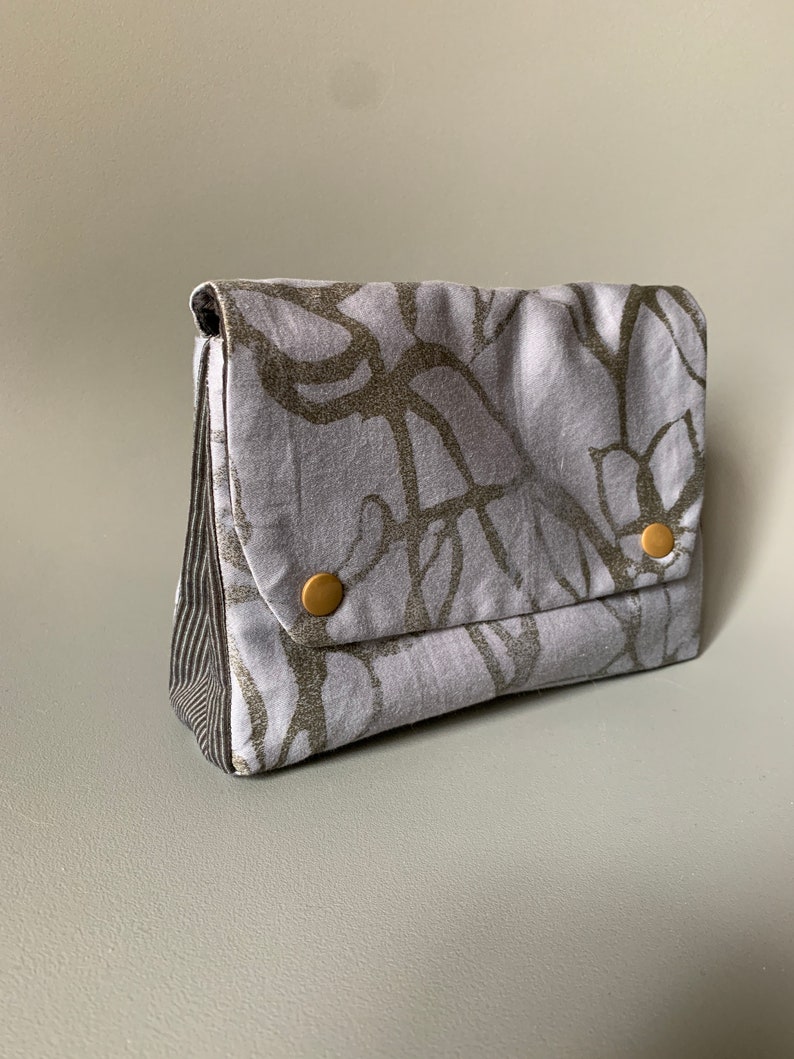 May include: A small, gray fabric pouch with a silver and black abstract pattern. The pouch has a flap closure and two gold buttons.