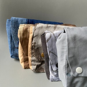 May include: Five fabric pouches in different patterns and colors. The pouches are lined up from left to right, with the blue denim pouch on the left and the gray pouch on the right. The pouches are all made of different fabrics, including denim, plaid, and solid colors.