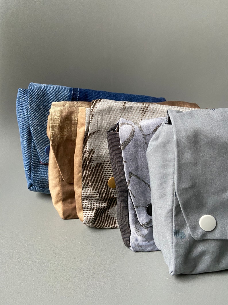 May include: Five fabric pouches in different patterns and colors. The pouches are lined up from left to right, with the blue denim pouch on the left and the gray pouch on the right. The pouches are all made of different fabrics, including denim, plaid, and solid colors.
