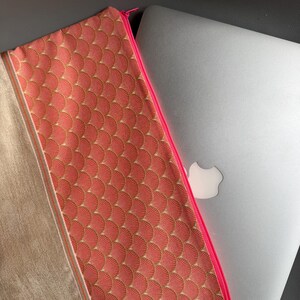 Peut inclure: Une pochette zippée corail avec un motif doré en forme d'éventail et une fermeture éclair rose vif. La pochette est à côté d'un ordinateur portable argenté. Le côté gauche de la pochette est de couleur beige clair.