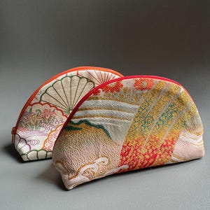 May include: Two half-moon shaped fabric pouches with red zippers. The pouch on the left has a white background with a green, pink, and orange floral pattern. The pouch on the right has a white background with a green, gold, and red geometric pattern.