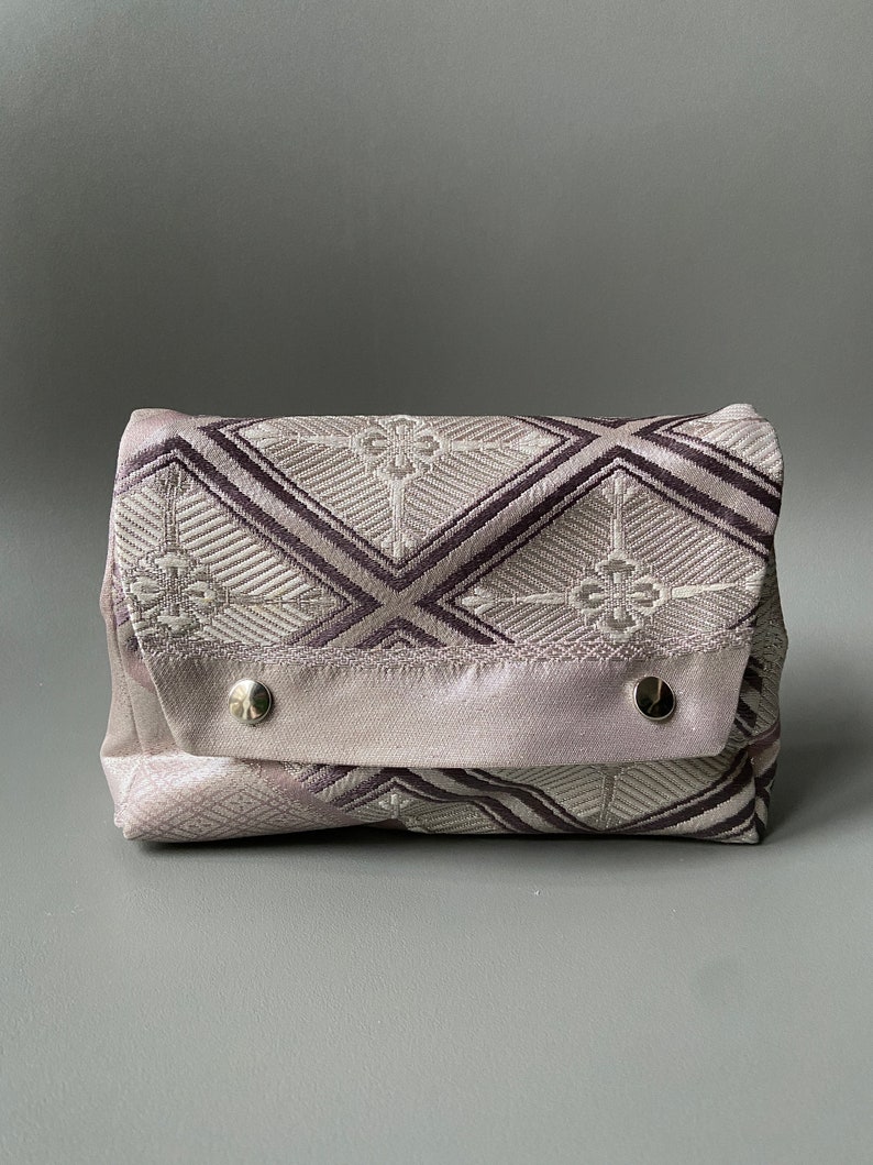 May include: A small, rectangular, gray and white patterned pouch with a flap closure and two silver snaps. The pouch is made of a soft, textured fabric.