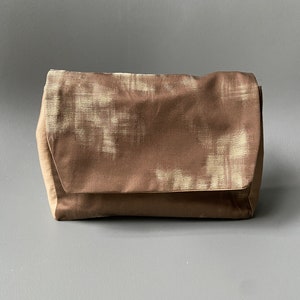 May include: A brown clutch purse with a white pattern. The purse has a flap closure and a soft, textured fabric.