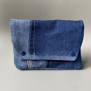 May include: A blue denim clutch purse with a flap closure and two blue buttons. The purse is made from different shades of denim and has a red stitching detail.
