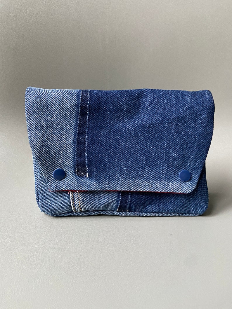 May include: A blue denim clutch purse with a flap closure and two blue buttons. The purse is made from different shades of denim and has a red stitching detail.