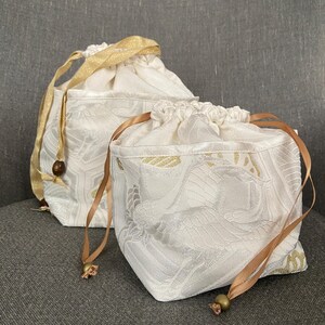 以下が含まれることがあります： 金色と茶色のリボン紐が付いた白い布製の巾着袋が2つ。袋にはさりげないエンボス加工が施されており、小さな物の収納に最適です。
