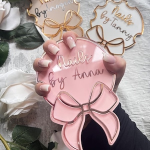 Può includere: Tre cartelli per salone di manicure in acrilico rosa e bianco con finiture dorate e un fiocco. I cartelli recitano "Nails by Gem", "Nails by Tammy" e "Nails by Anna".