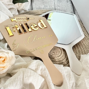 May include: A beige hand mirror with a gold inscription that reads "Filled by Clinic co @CLINIXCOMPANY". The mirror has a white handle and a gold syringe icon.