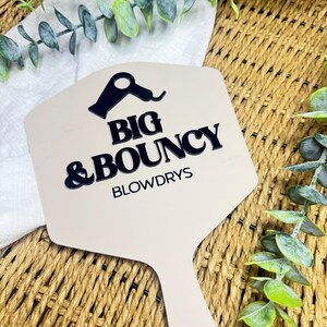May include: A beige hand mirror with a black graphic of a hair dryer and the black text BIG & BOUNCY BLOWDRYS printed on the surface. The mirror has a hexagonal head and a handle, featuring a clean, flat design with bold lettering.