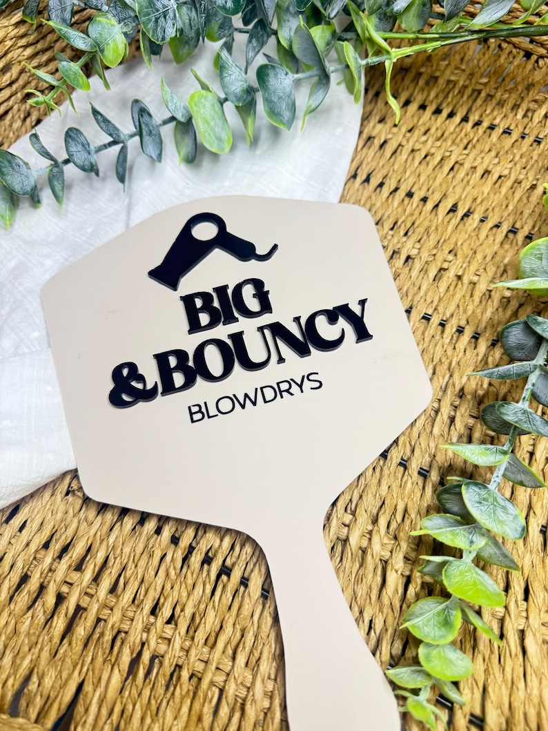 May include: A beige hand mirror with a black graphic of a hair dryer and the black text BIG & BOUNCY BLOWDRYS printed on the surface. The mirror has a hexagonal head and a handle, featuring a clean, flat design with bold lettering.
