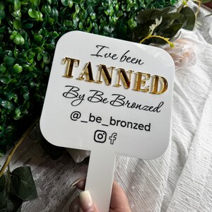 May include: White sign with black text that reads "I've been tanned by Be Bronzed @be_bronzed" with a gold Instagram and Facebook icon. The sign is attached to a white stick.