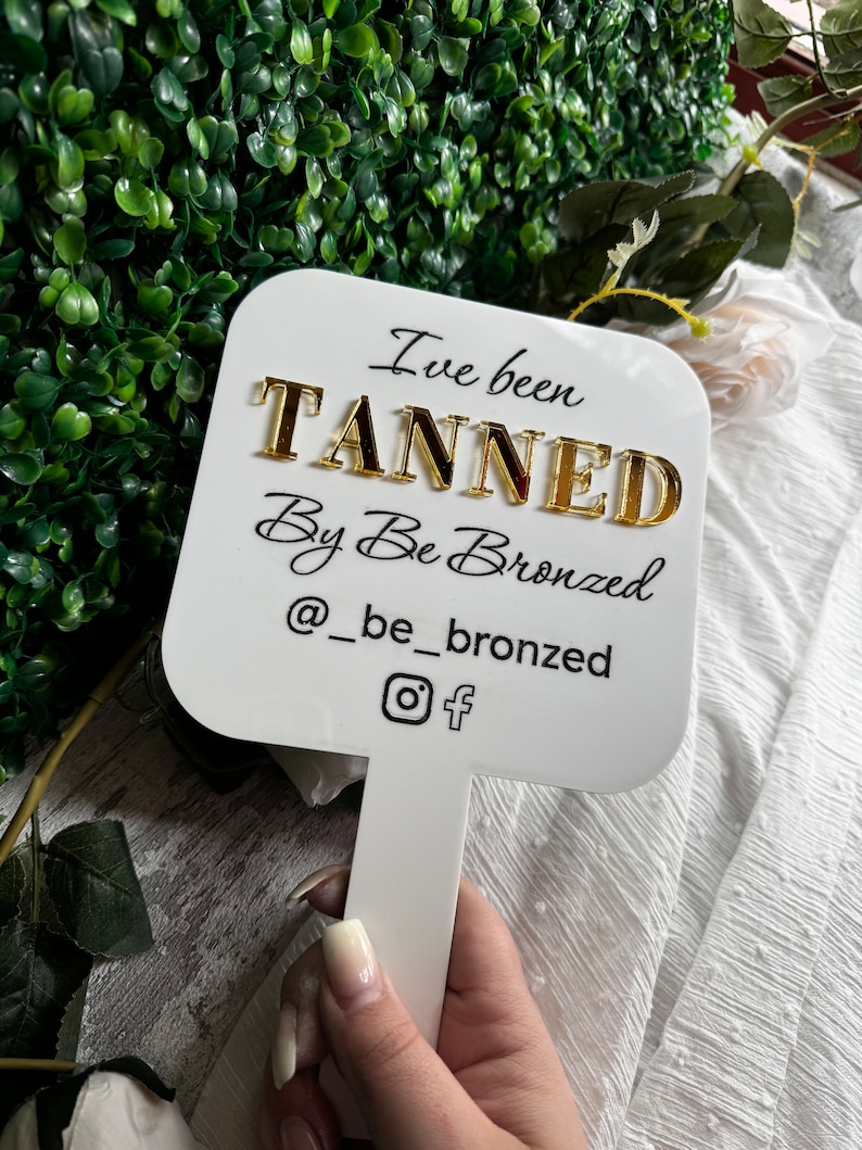May include: White sign with black text that reads "I've been tanned by Be Bronzed @be_bronzed" with a gold Instagram and Facebook icon. The sign is attached to a white stick.