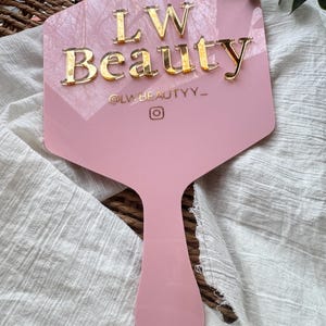 May include: A light pink, hexagonal handheld mirror featuring the gold, raised text LW Beauty, the handle @LWBEAUTYY_, and an Instagram icon. The mirror has a smooth, glossy finish and a long, tapered handle extending from the base of the hexagon.