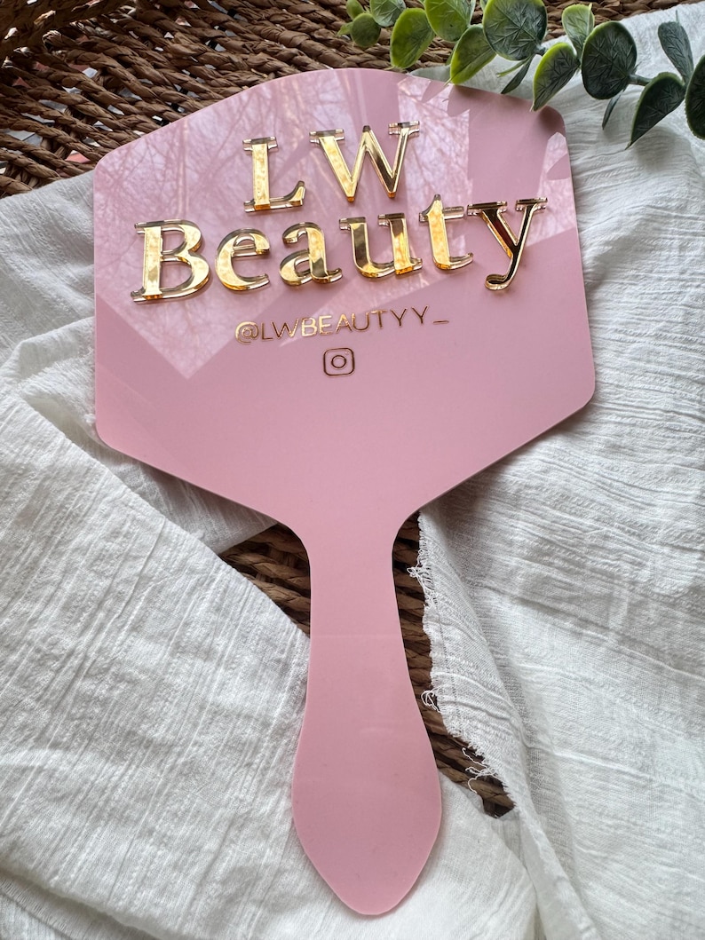 May include: A light pink, hexagonal handheld mirror featuring the gold, raised text LW Beauty, the handle @LWBEAUTYY_, and an Instagram icon. The mirror has a smooth, glossy finish and a long, tapered handle extending from the base of the hexagon.