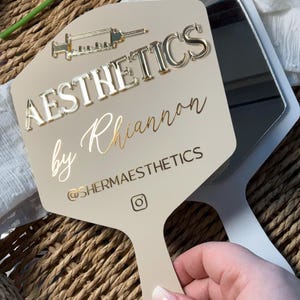 May include: A beige handheld mirror featuring a gold syringe icon and the text AESTHETICS by Rhiannon with the handle @SHERMAESTHETICS and an Instagram logo. The mirror has a smooth, flat surface with raised gold lettering and a reflective glass back.
