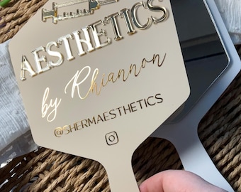 May include: A beige handheld mirror featuring a gold syringe icon and the text AESTHETICS by Rhiannon with the handle @SHERMAESTHETICS and an Instagram logo. The mirror has a smooth, flat surface with raised gold lettering and a reflective glass back.
