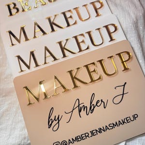 K&ouml;nnte beinhalten: Ein Stapel aus vier rechteckigen Schildern mit abgerundeten Ecken. Die oberen drei sind wei&szlig; mit goldenen, verspiegelten 3D-Buchstaben, die BEAUTY und MAKEUP lesen. Das untere Schild ist beige mit goldenen 3D-Buchstaben MAKEUP, Amber J und @AMBERJENNASMAKEUP.