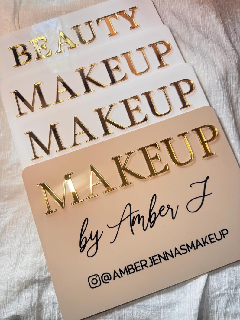K&ouml;nnte beinhalten: Ein Stapel aus vier rechteckigen Schildern mit abgerundeten Ecken. Die oberen drei sind wei&szlig; mit goldenen, verspiegelten 3D-Buchstaben, die BEAUTY und MAKEUP lesen. Das untere Schild ist beige mit goldenen 3D-Buchstaben MAKEUP, Amber J und @AMBERJENNASMAKEUP.