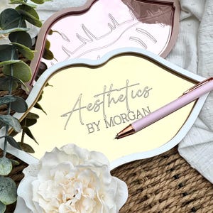 May include: Gold and rose gold lip-shaped trays with a white border. The gold tray has the text "Aesthetics by Morgan" written on it. A pink pen is lying on the tray.