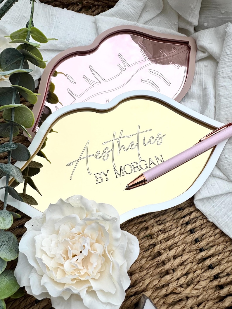 May include: Gold and rose gold lip-shaped trays with a white border. The gold tray has the text "Aesthetics by Morgan" written on it. A pink pen is lying on the tray.